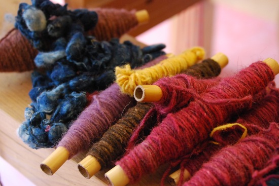 A close-up of a pile of bobbins of wool, in a variety of colours. On the right are reds and browns, and on the left are blues; there's also a small bundle of yellow yarn on top of teh pile. 
